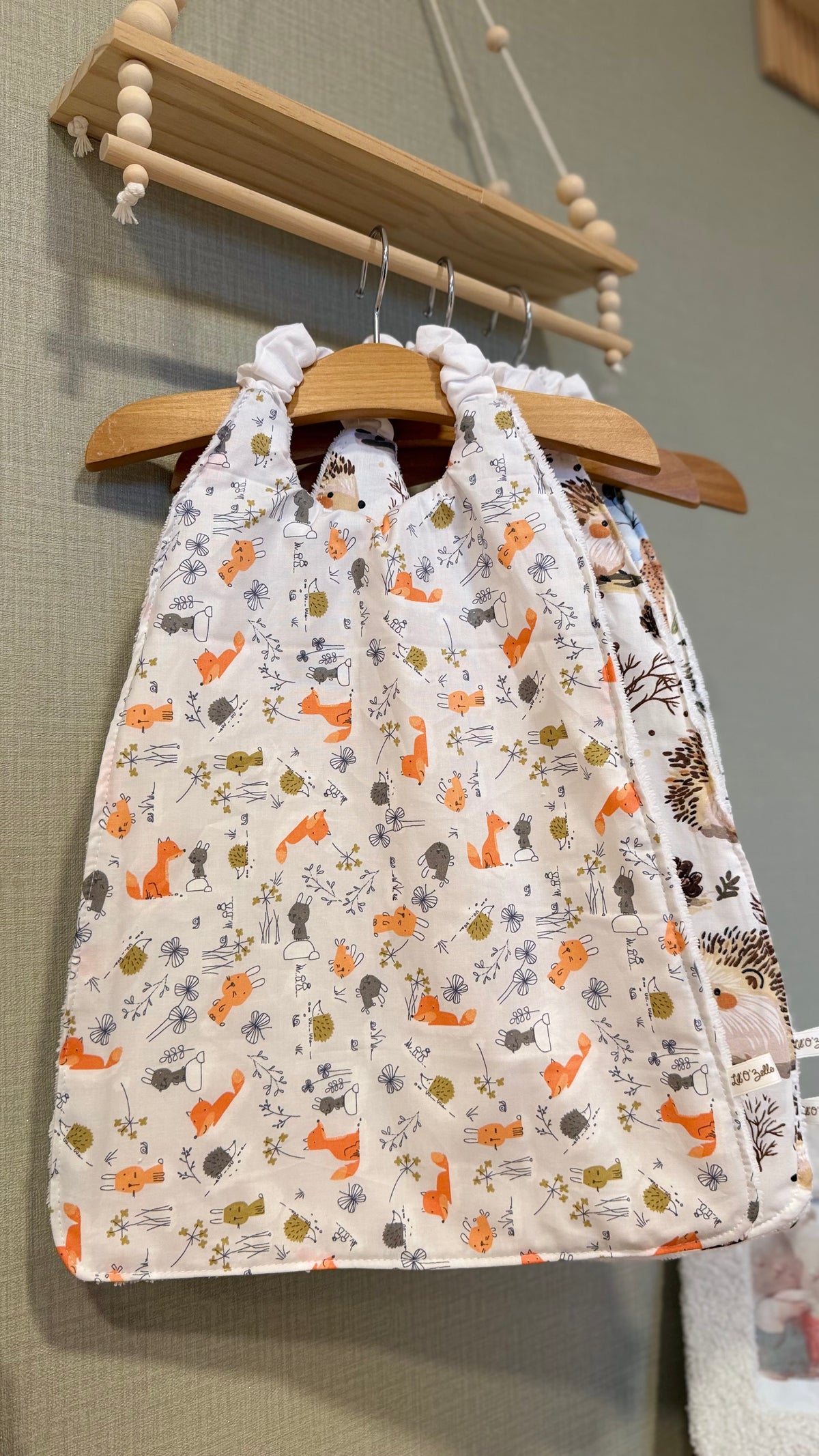 Grand bavoir maternelle Zouzou motif renards et fleurs, vue de face sur un cintre en bois